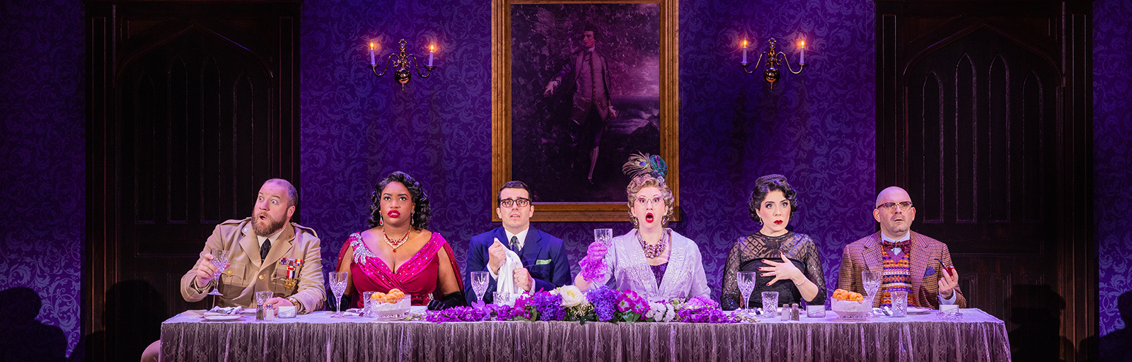 Six actors in formal attire sit all on the same side behind a long fancy dinner table. Each actor is staring forward into the distance with a shocked and perplexed look.